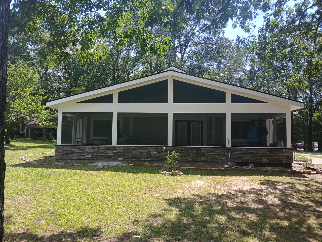 A single-story house with a stone foundation and screened-in front porch, surrounded by grass and trees on a sunny day.