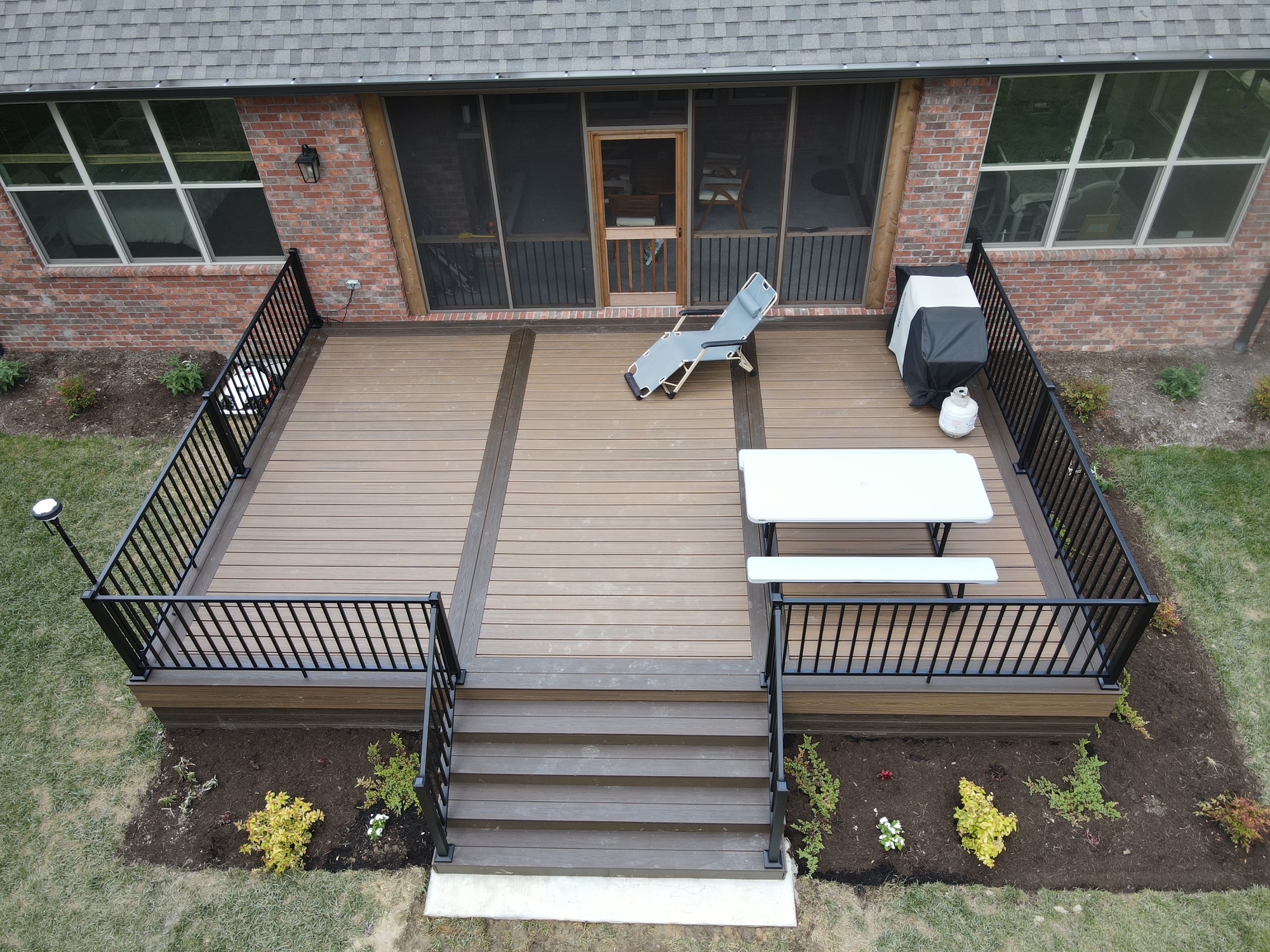 Aerial view of a wooden backyard deck with black railings, steps, a picnic table, a lounge chair, a grill, and landscaped garden beds around the perimeter.