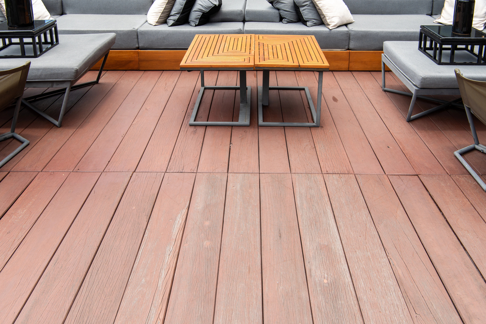 Outdoor seating area with gray cushioned benches and chairs, a wooden deck floor, and two square wooden tables with metal frames placed in the center.