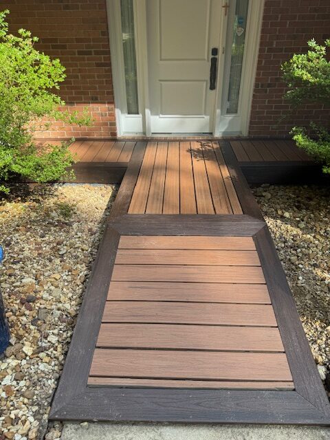A wooden ramp with dark trim leads to a white front door of a brick house, surrounded by shrubs and decorative rocks.