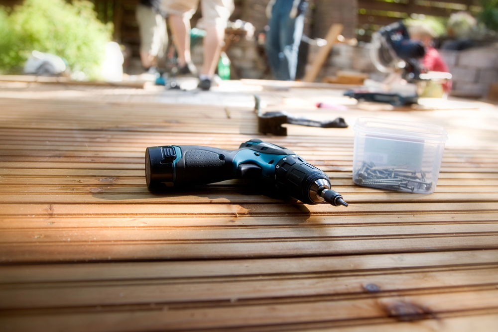 A cordless power drill and a plastic box of screws are placed on a wooden deck, with people working in the blurred background.
