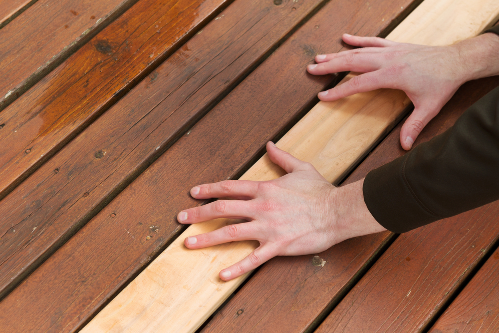Two hands position a new light-colored wooden plank among older, darker stained wooden boards in a deck or floor repair setting.