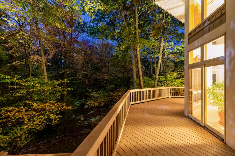 A wooden deck outside a house overlooks a forested area at dusk, with warm interior lights visible through large windows.