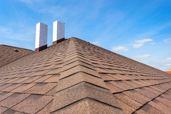 A close-up view of a sloped roof with brown asphalt shingles and two white rectangular chimneys against a blue sky.