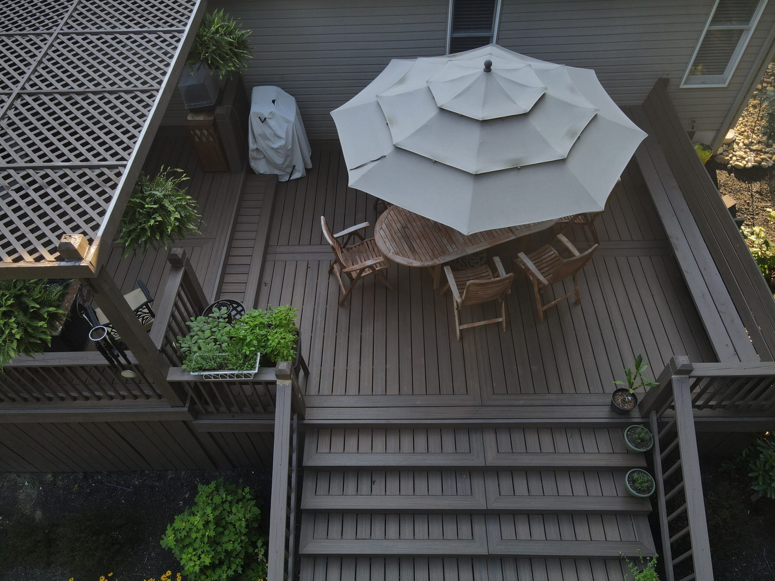 A wooden deck with an outdoor dining table, four chairs, a large white umbrella, potted plants, and a barbecue grill.